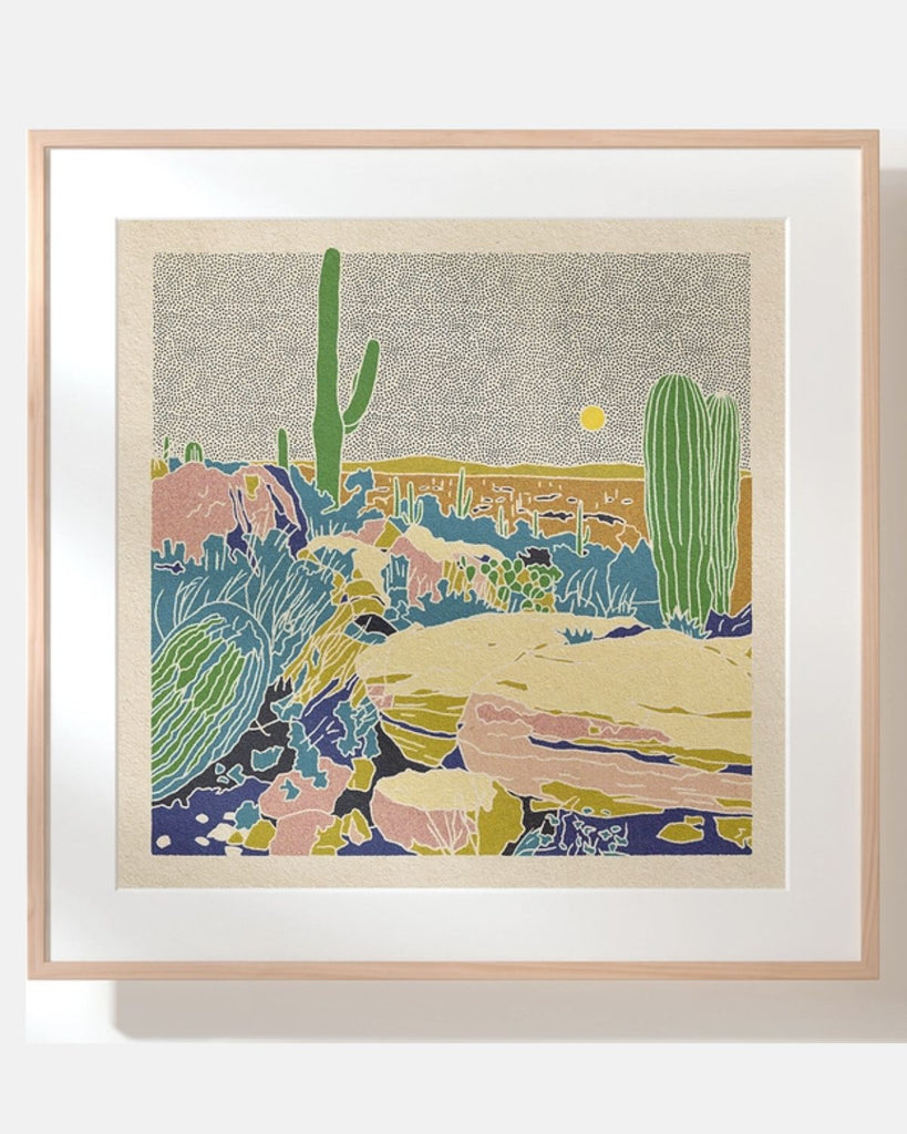 The Desertscape print by Colorado artist,in a wooden frame. The print shows cactus and rocks in the foreground and flat hills in the background. The sky is filled with small black dots with a large yellow dotted sun. The cactuses are hughs of green and blue and the rocks are pinks, yellows, and tans. The outlines are white giving helping to define each colorful shape.