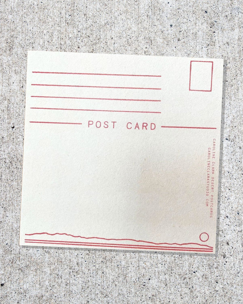 The back of the postcard shown on a cement surface. The back has red lines for an address and stamp, and it reads 'postcard' below it. On the bottom is a thin couple of lines that make out a horizon with a small circle for a sun. 