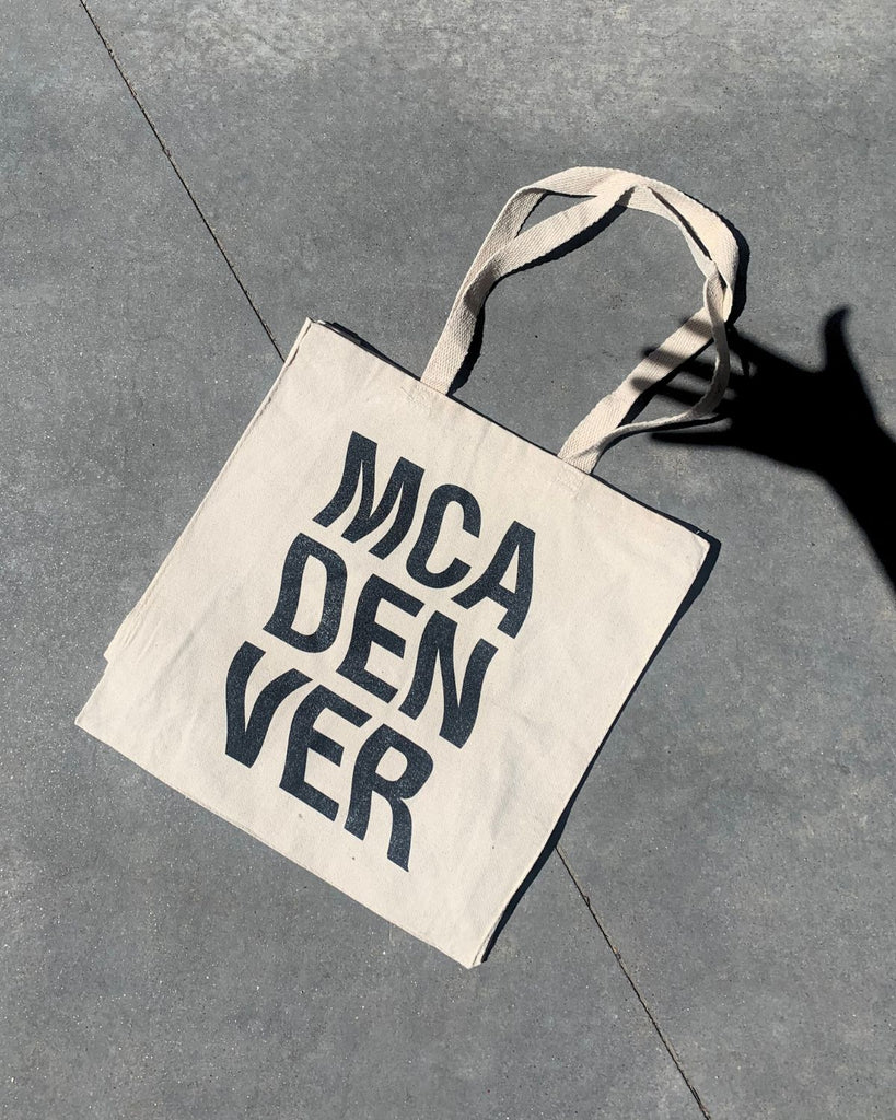 MCA Denver Wavy Tote. Natural canvas tote with black bold squiggle letters that read 'MCA' on top, 'DEN', in the middle, and 'VER' on the bottom. 