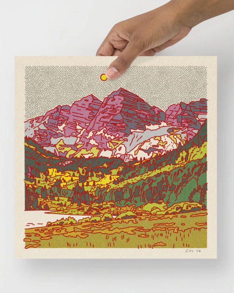 A hand holding the desert mountain print by Colorado artist, Caroline Clark. The art print shows a mountain scene with water and grass in the foreground, trees in the middle ground, and a rocky mountain in the background. The sky is spotted with black dots and a bigger yellow dot for the sun. Shades of light and dark green color the grass and trees while the mountain is shades of pinks and purples. The outline is in red helping to define each colorful shape.
