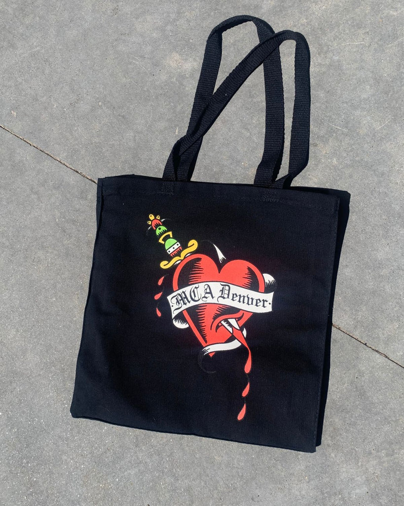 MCA Denver Heart Tote. Tote is black with a red heart pierced with a green and yellow sword. A white ribbon that reads 'MCA Denver' is wrapped around the heart. Art is done in a traditional tattoo style.  