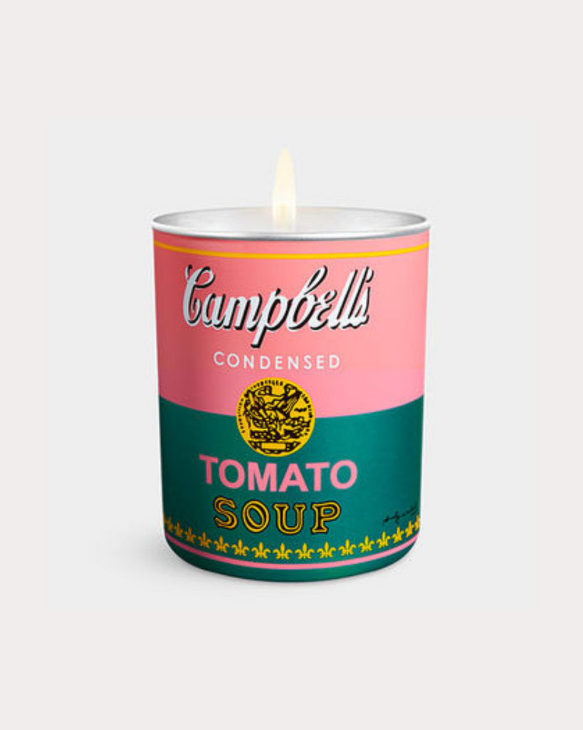 Candle has a lit wick and is against a white backdrop. The candle resembles the famous Warhol soup cans artwork and has pink on the top half and green on the bottom half. The text on the candle reads 'Campbell's condensed tomato soup' 