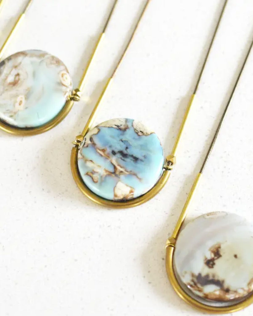 Arctic Terra Agate stone necklaces show flat against a white surface. The stones are cradled in a brass half-circle. 