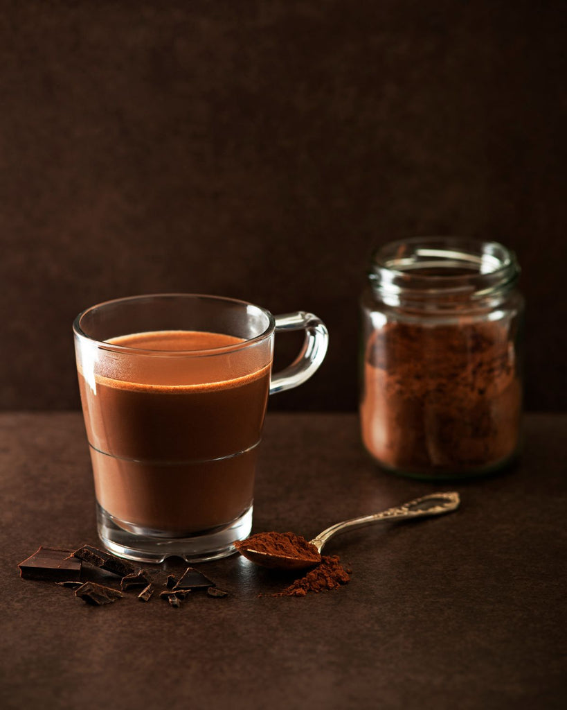Mexican Drinking Chocolate shown in a clear mug with coco nibs and powder behind it. 