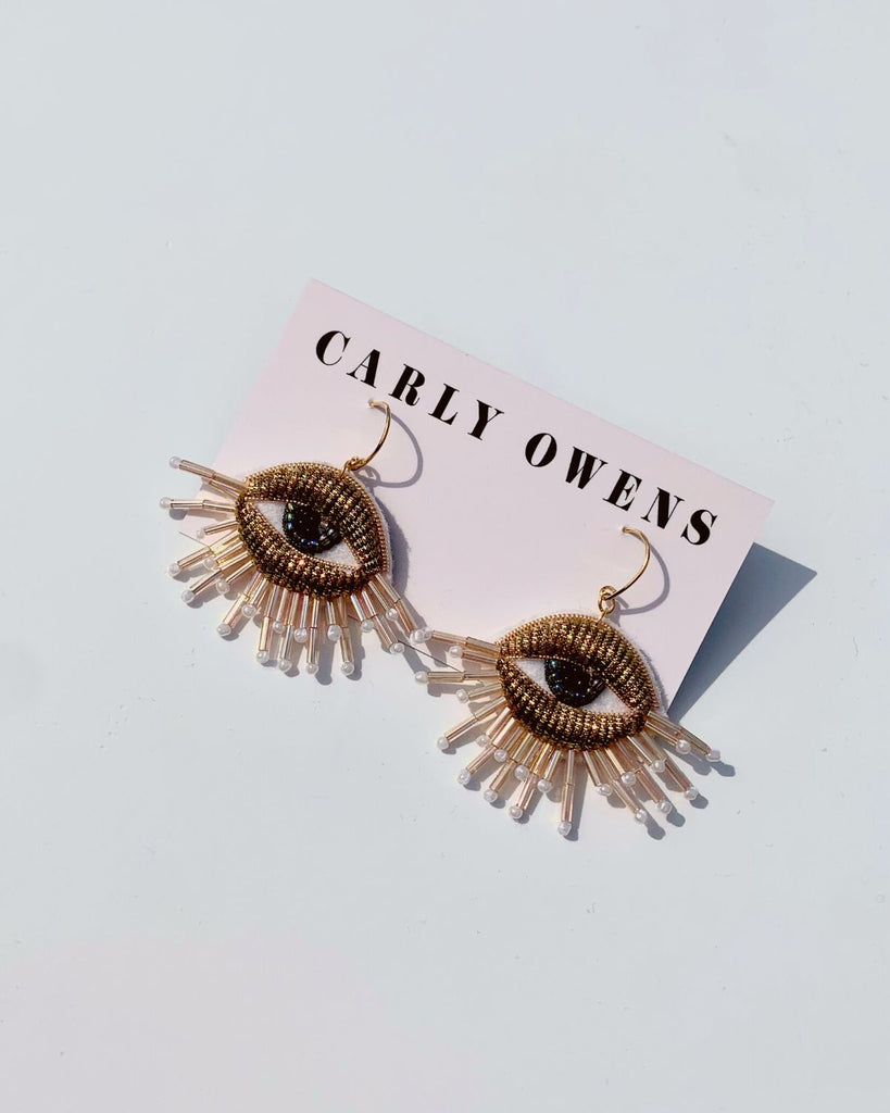Embroidered eye earrings shown on their branded paper backing that reads 'Carly Owens' across the top. The Eye earrings have gold beaded eyelids, dark blue beaded eyes, and light pink metallic beaded eyelashes along the bottom lid. 