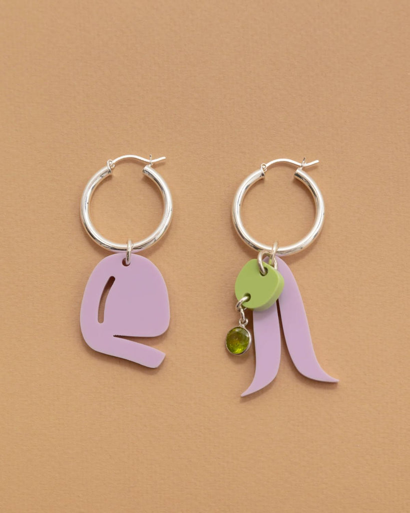 The Lilac - Sour Apple earrings are shown on a tan paper surface. The hoops are a thick silver with acrylic charms attached. Both earrings have a lilac abstract shape charm but one of the earrings has a pastel green charm and gem added. 