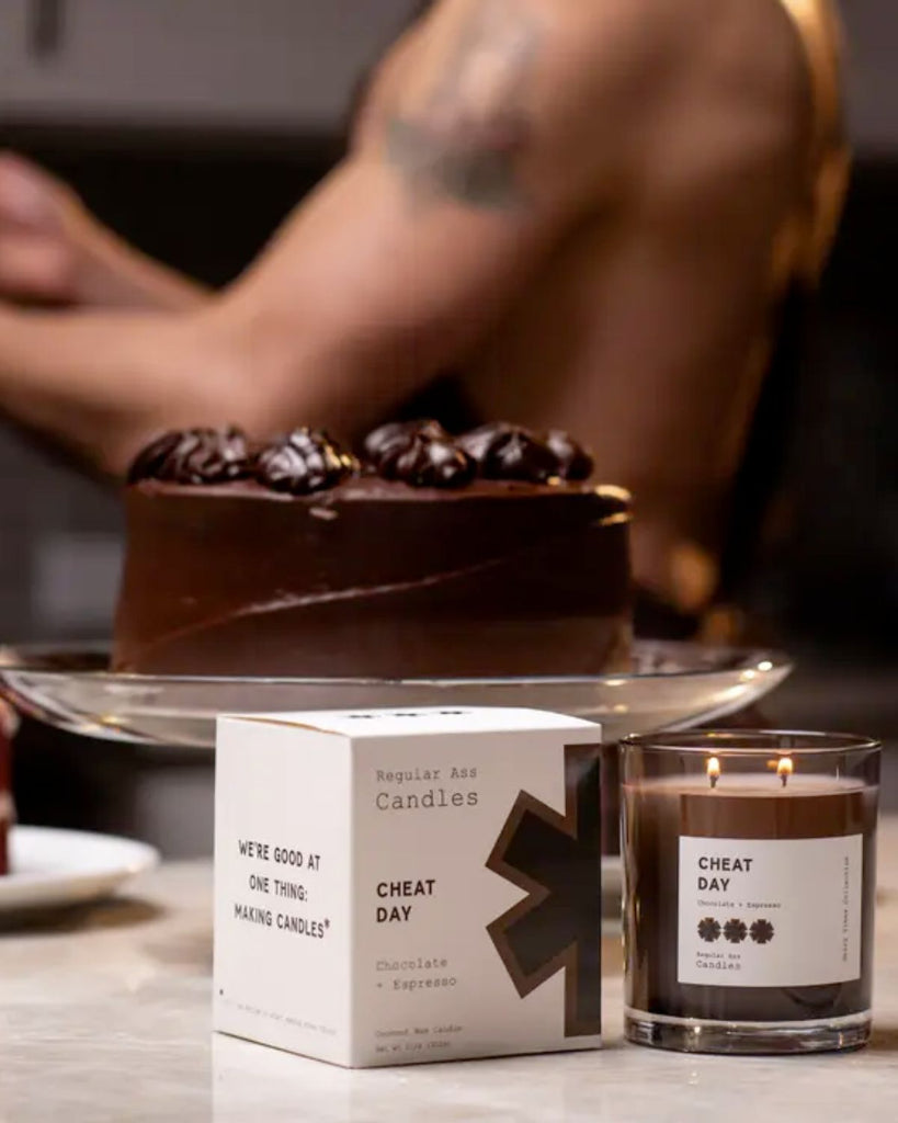 Candle is lit on a counter with a chocolate cake and a shirtless man in an apron in the background. 