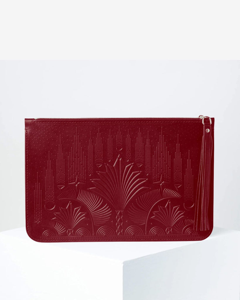 The clutch sits on a pedestal against a white backdrop. The clutch is a deep burgundy color and has a geometric floral embossed design on the front and back. 