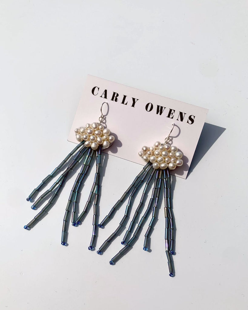 Cloud earrings shown with their paper backing that reads Carly Owens on a white surface. The earrings have silver hook backings attached to pearl beaded cloud shapes with blue bead raindrops hanging from them.