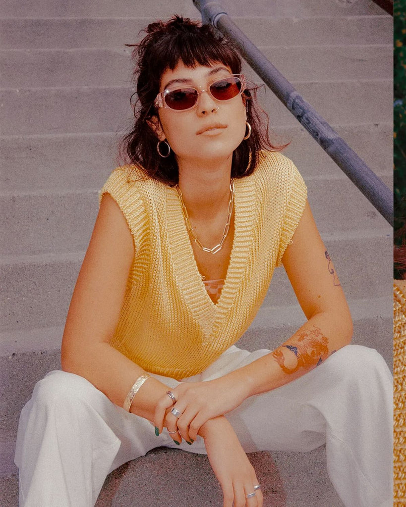 The Funk Punk sunglasses on a female presenting model. The model is outside sitting on stairs wearing a yellow sweater vest with white pants. 