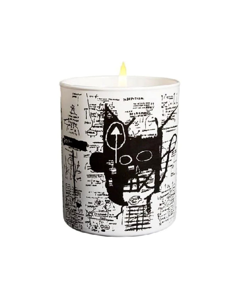 The candle shown with a lit wick against a white backdrop. The Basquiat artwork has black scribbles and drawings and a focal point of a face colored in black.