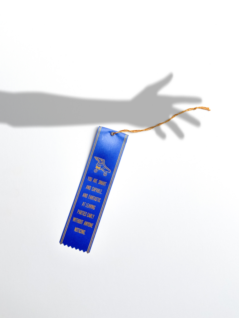 The blue ribbon with gold text is shown on a white surface. A shadow of a hand is shown over the ribbon. 