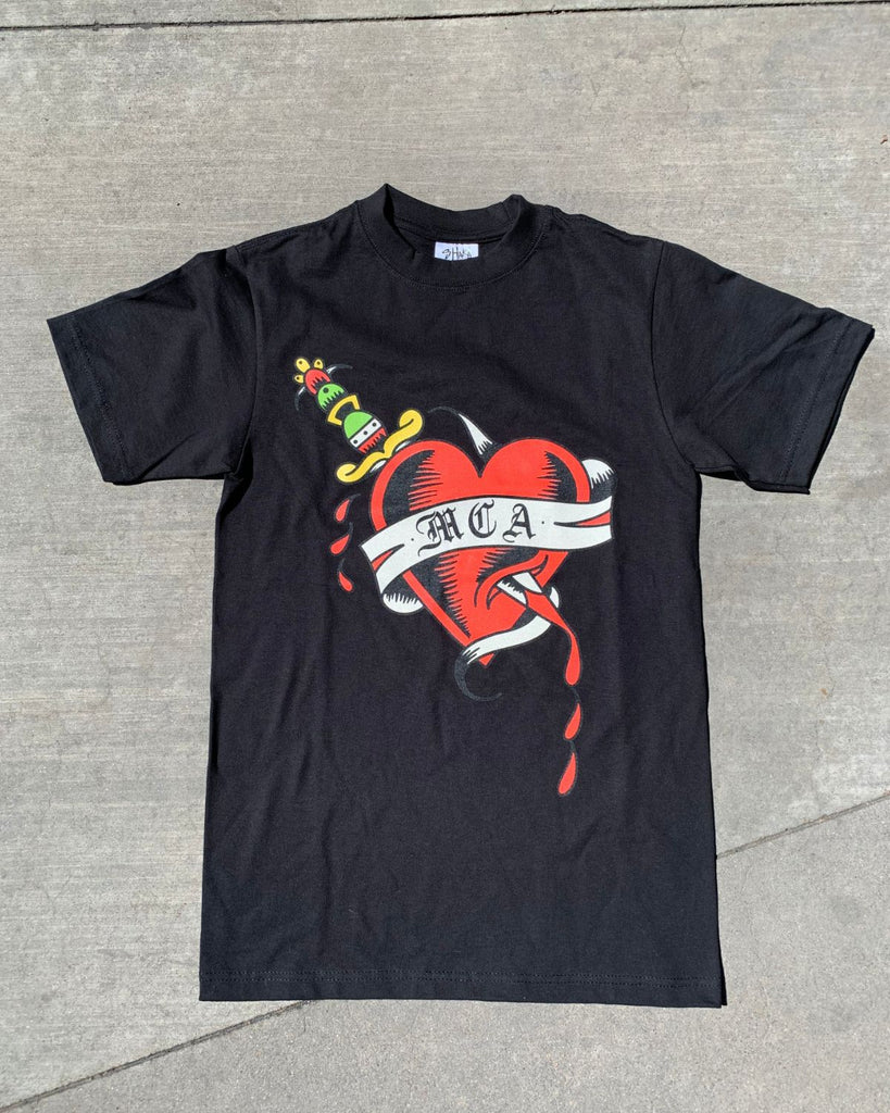 The MCA Denver Heart T-shirt is black with a traditional tattoo styled heart and dagger. A ribbon lays on top of the heart and reads in old english style font, "MCA". 