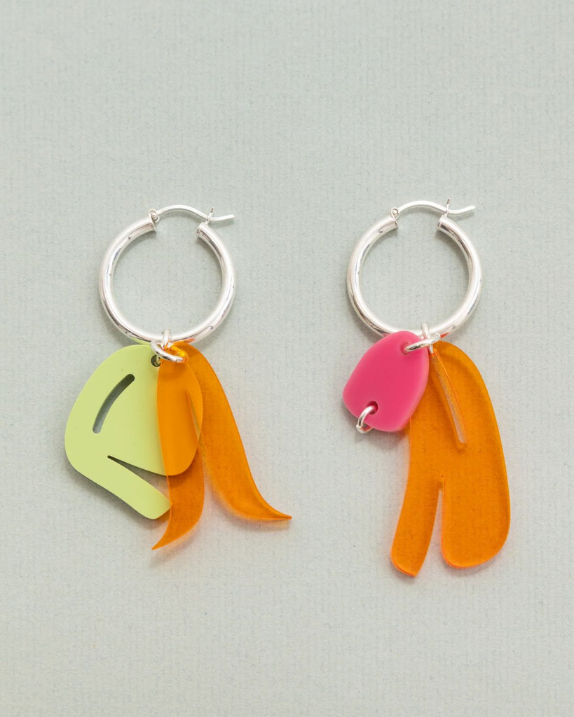 The Orange Multi earrings are shown against an off-white surface. The earrings have a thick silver hoop with abstract acrylic charms linked on. Both earrings have translucent orange charms but one hoop has an opaque green charm with it and the other hoop has an opaque pink charm. 