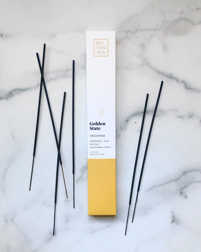Incense packaging with incense sticks beside it. The packaging is white with mustard yellow on the bottom. 