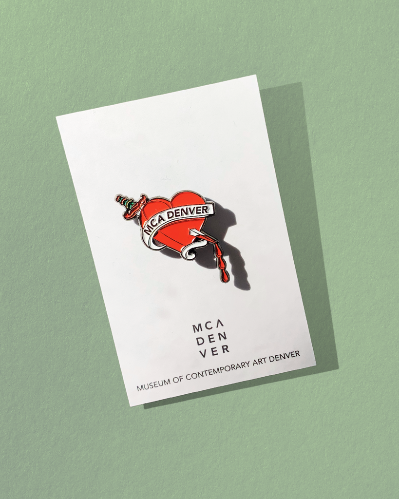 MCA Denver Heart Pin on the cardstock backing on top of a mint green paper surface. 