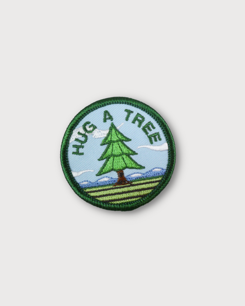 The Hug a Tree embroidered patch is made right here in Denver! The patch is circular and shows a single tree in the middle. Around the tree in a half circle reads 'hug a tree' in green embroidered letters. 