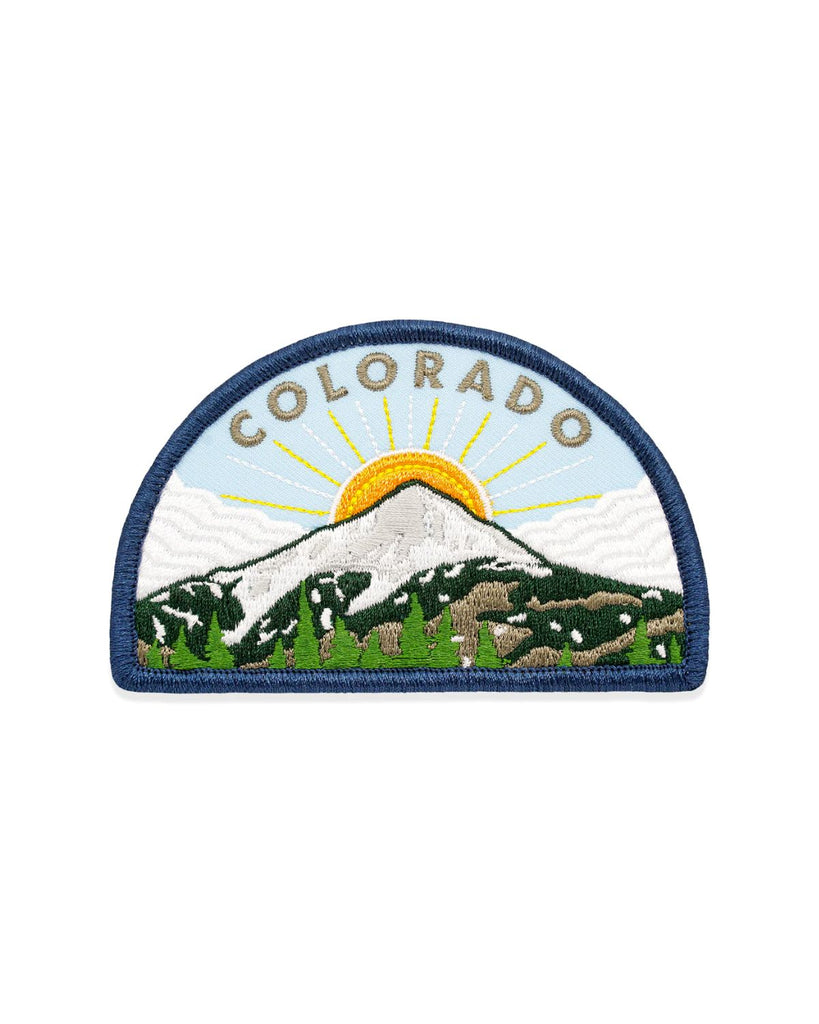 The Colorado Mountain Patch is made right here in Denver. The patch is a half circle shape with a snow-capped mountain in the center. A sun emerges from the mountain peek and has long yellow rays extended from it. The sky is light blue with wavy white clouds. Embroidered grey letters read 'Colorado' across the top. 