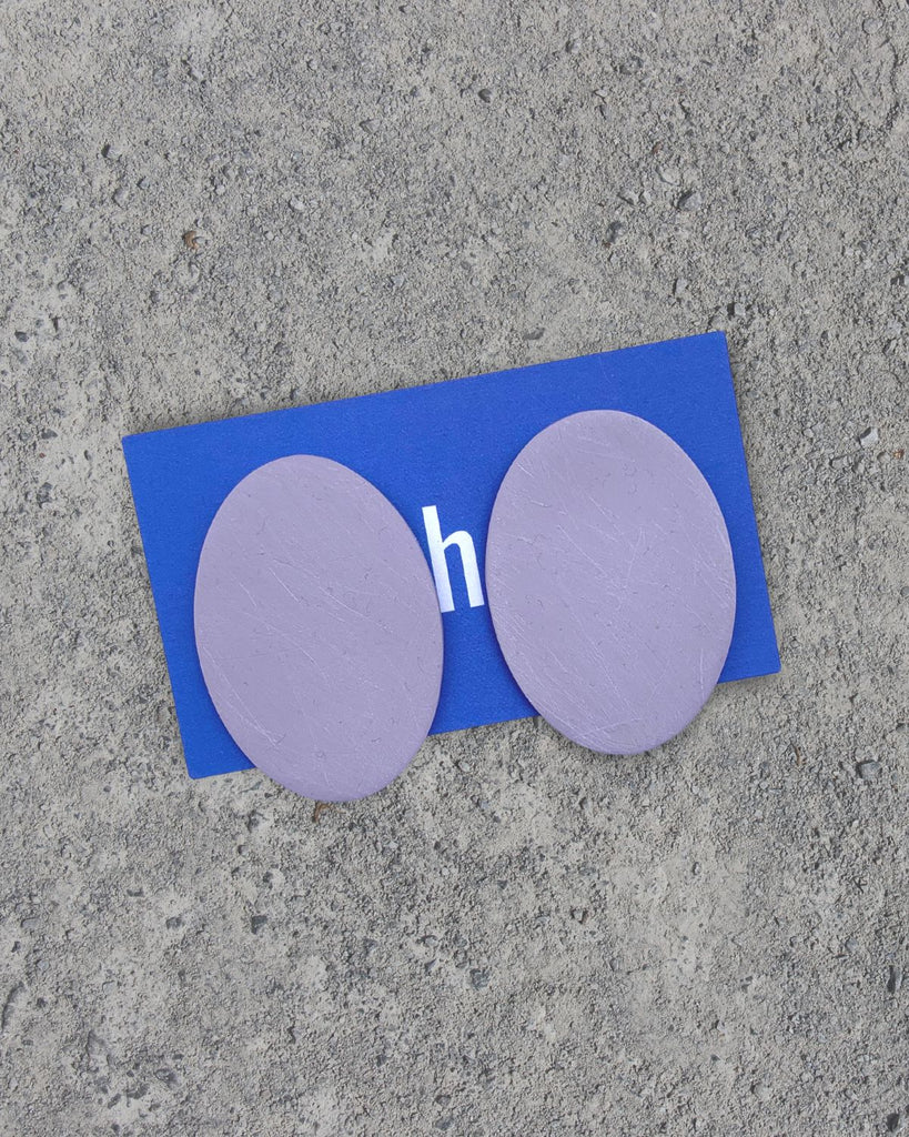 The Lavender earrings are laid on a cement surface. The earrings are oval shaped and have a slight brushed texture on the surface. 