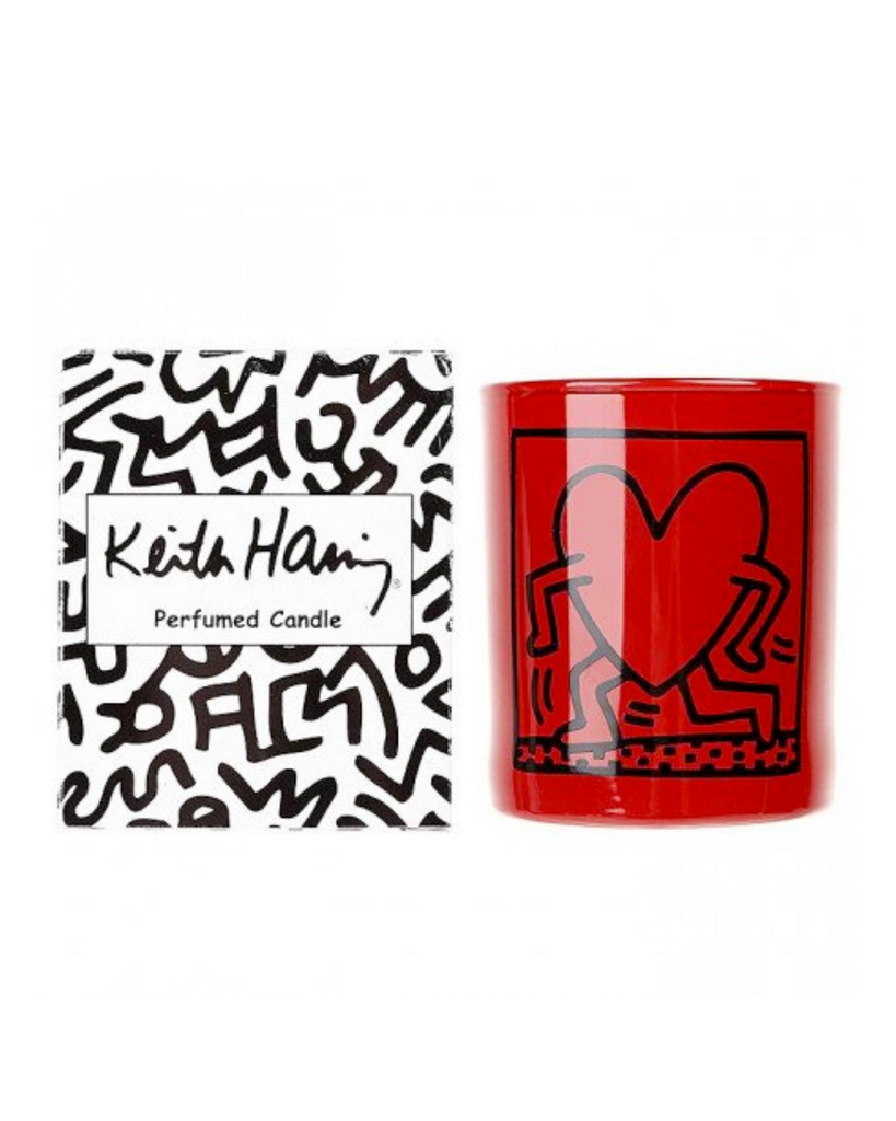Candle shown with the box it comes with against a white backdrop. The box is on the left and is covered in black and white Haring doodles. The candle on the right is red with Harings famous running heart artwork on the front. The drawing is in black. 
