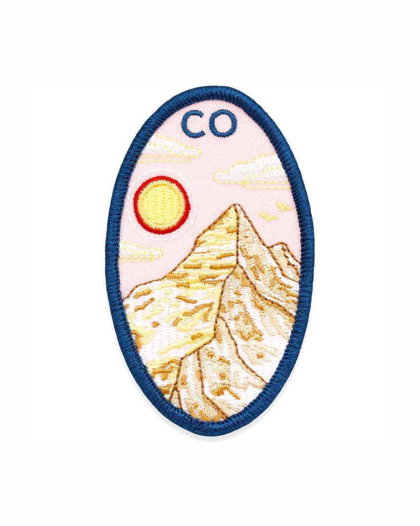 The Colorado San Juans patch is made right here in Denver! The patch is a long vertical oval shape with a navy blue border. Inside the border is an embroidered mountain in gold and yellow colors. A pink sky with a glowing yellow and red sun is behind it. On top of the scene are embroidered letters that read 'CO'.  