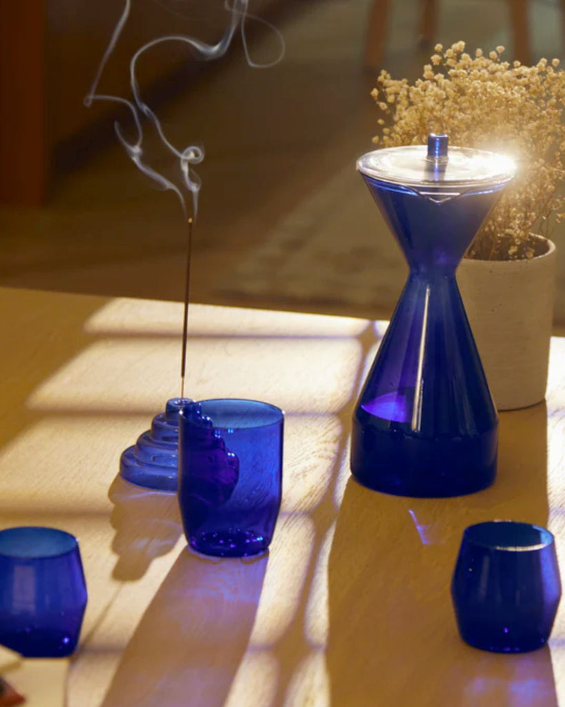 The Incense holder in use on a light wooden table. Shown with three cobalt glasses in the foreground and the Cobalt Carafe on the right. Smoke from the incense holder travels up to the left.