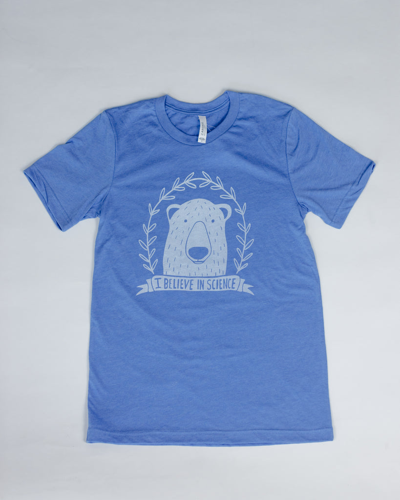 The t-shirt laid flat on a white surface. The shirt is blue and has an illustration of a polar bear surrounded by a laurel wreath with a ribbon on the bottom that reads 'I believe in science'.